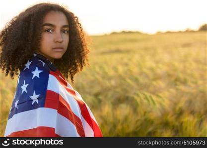 Sad depressed mixed race African American girl teenager female young woman with tears in her eyes in a field of wheat or barley crops holding and wrapped in USA stars and stripes flag in golden sunset evening sunshine