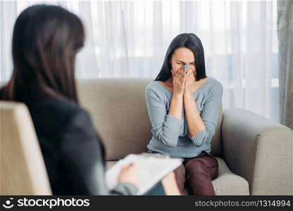 Sad female patient sitting on sofa at psychotherapist reception. Female doctor writes notes in notepad, professional psychology support. Female patient sitting on sofa at psychotherapist