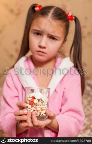 Sad little girl with glass full of tablets and pills in her hands.