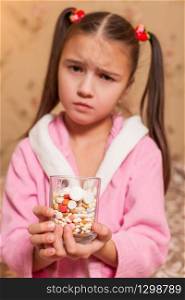 Sad little girl with glass full of tablets and pills in her hands.