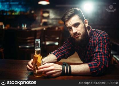 Sad man sitting at the bar counter and holds the bottle with alcohol beverage in hands. Male person in pub, alcoholism, alcohol addiction, drunkenness. Man sitting at the bar counter, alcohol addiction