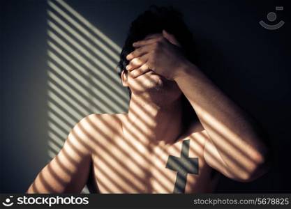 Sad naked man sitting by the window with shadows from blinds on his face