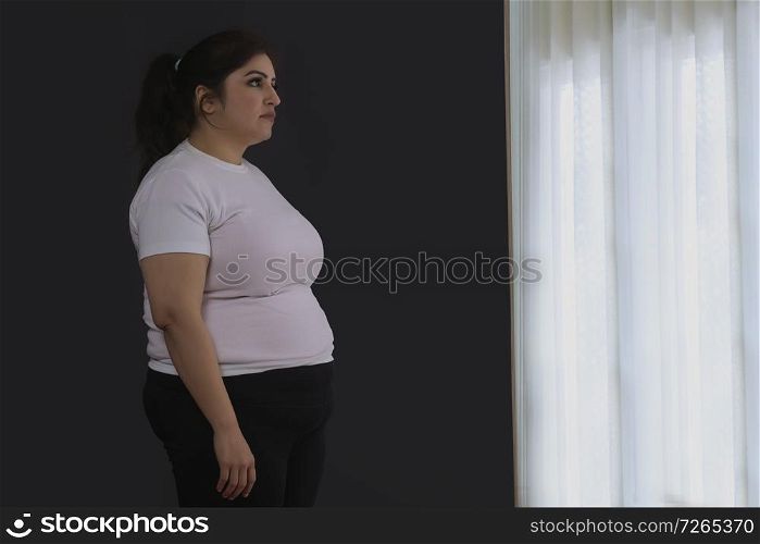Sad obese woman looks while standing by the window