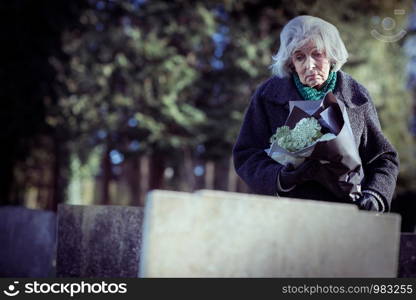 Sad Senior Woman With Flowers Standing By Grave