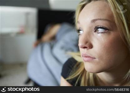 Sad Teenage Girl Sitting In Bedroom Whilst Boyfriend Sleeps