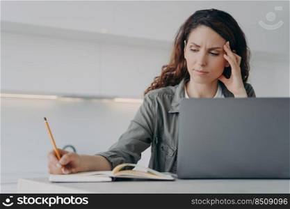 Sad tired freelancer is working at home. Exhausted european woman is sitting in front of laptop and taking notes. Overworked girl has got a headache. Concept of remote work stress.. Sad tired freelancer working at home. Exhausted woman sitting in front of laptop and taking notes.