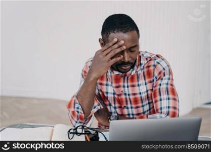 Sad tired online teacher or businessman. Afro man has online conference on laptop or working on project late. Tutor is exhausted from work. Remote study, distance learning. Deadline concept.. Sad tired online teacher or businessman. Remote study or work, distance learning. Deadline concept.