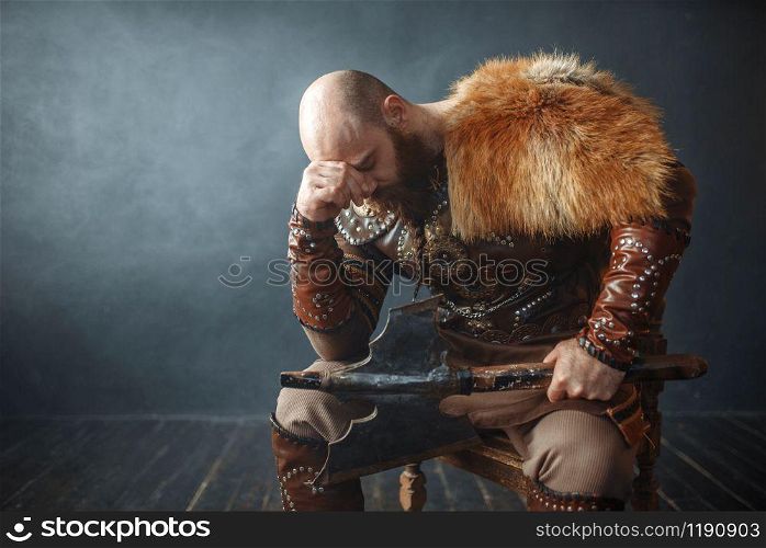 Sad viking with axe dressed in traditional nordic clothes sitting on chair, barbarian image. Ancient warrior in smoke on dark background. Sad viking with axe sitting on chair