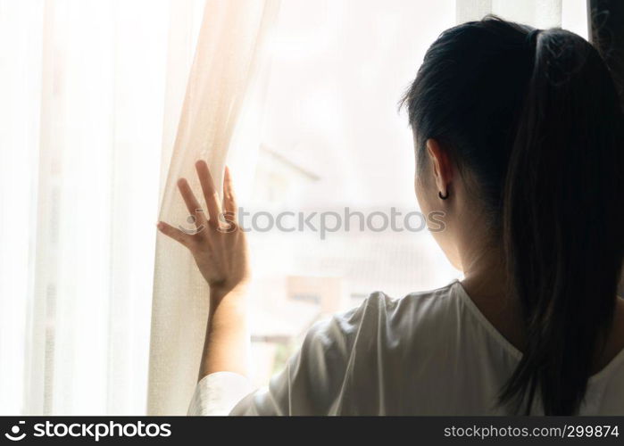 sad woman holding the curtains open to look out of a large light window at home, interior