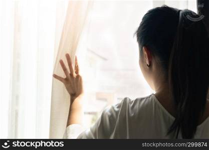 sad woman holding the curtains open to look out of a large light window at home, interior