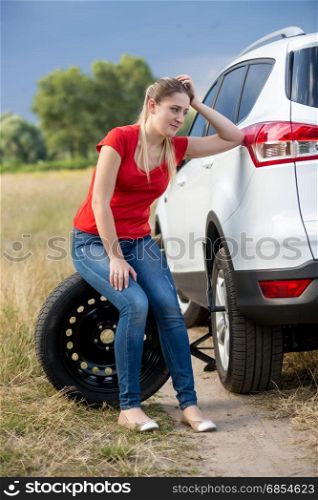 Sad woman sitting on tire and leaning on car at countryside road
