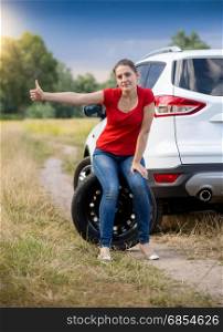 Sad woman sitting on wheel at broken car in filed and hitchhiking
