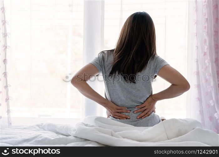 Sad young Asian woman touching back feeling backache morning discomfort low lumbar muscular kidney pain sit on bed after bad sleep waking up on uncomfortable mattress bending. Concept of woman stretching suffering from sudden back pain.