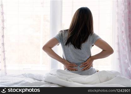 Sad young Asian woman touching back feeling backache morning discomfort low lumbar muscular kidney pain sit on bed after bad sleep waking up on uncomfortable mattress bending. Concept of woman stretching suffering from sudden back pain.