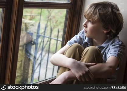 Sad young boy sitting, thinking and looking out of rain covered window
