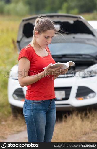 Sad young woman reading owner manual at broken car in meadow