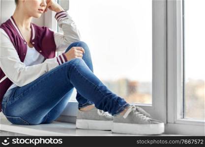 sadness and people concept - sad teenage girl sitting on window sill. sad teenage girl sitting on window sill