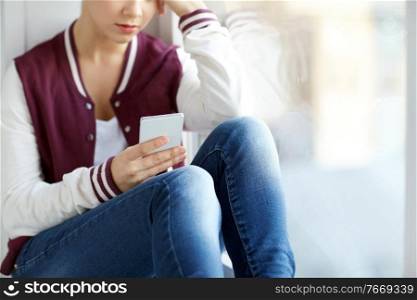 sadness and people concept - sad teenage girl with smartphone sitting on window sill. teenage girl with smartphone sitting on windowsill