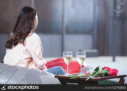 Sadness love in ending of relationship concept,Two glasses of wine and brunch of red roses on table wait for boyfriend with blurred broken heart young asian woman sitting in back side in background.