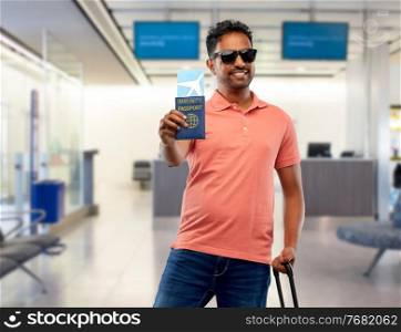 safe travel, tourism and health care concept - happy smiling young indian man with air ticket and immunity passport over airport background. happy man with air ticket and immunity passport
