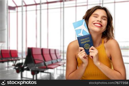 safe travel, tourism and health care concept - happy smiling young woman with air ticket and immunity passport over airport background. happy woman with air ticket and immunity passport