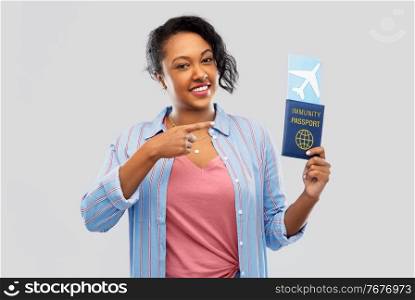 safe travel, tourism and health care concept - happy young african american woman with air ticket and passport over grey background. happy woman with air ticket and immunity passport