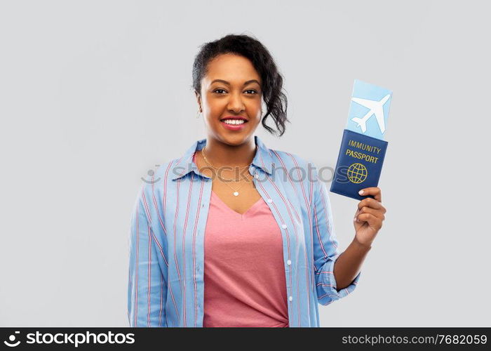 safe travel, tourism and health care concept - happy young african american woman with air ticket and passport over grey background. happy woman with air ticket and immunity passport