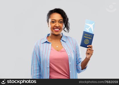 safe travel, tourism and health care concept - happy young african american woman with air ticket and passport over grey background. happy woman with air ticket and immunity passport