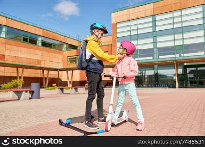 safety, childhood and care concept - happy school boy with backpack fastening girl&rsquo;s helmet for scooter riding. school boy fastening girl&rsquo;s helmet for scooter