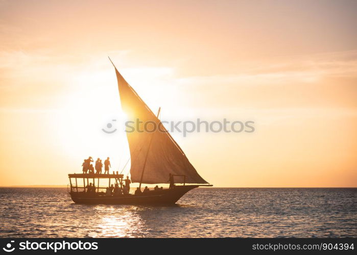 Sailboat in ocean at sunset in summer. Tropical landscape with silhouette of the people in boat, orange sky with gold sunlight. Yacht on the water in the evening. Travel in Zanzibar, Africa. Vacation