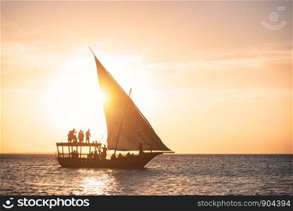 Sailboat in ocean at sunset in summer. Tropical landscape with silhouette of the people in boat, orange sky with gold sunlight. Yacht on the water in the evening. Travel in Zanzibar, Africa. Vacation
