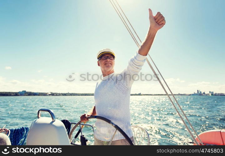 sailing, age, tourism, travel and people concept - happy senior man in captain hat on steering wheel and showing thumbs up sail boat or yacht floating in sea