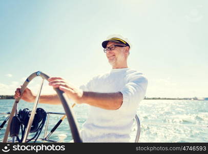 sailing, age, tourism, travel and people concept - happy senior man in captain hat on steering wheel and navigating sail boat or yacht floating in sea