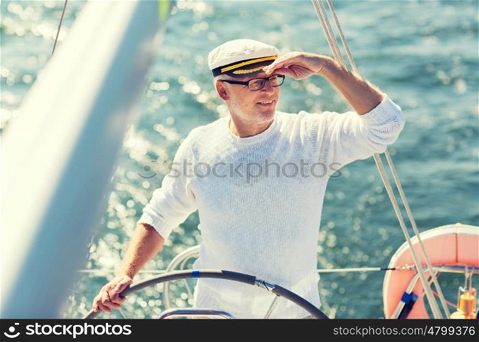 sailing, age, tourism, travel and people concept - happy senior man in captain hat on steering wheel and navigating sail boat or yacht floating in sea