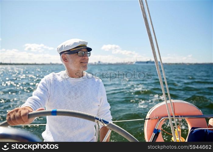 sailing, age, tourism, travel and people concept - happy senior man in captain hat on steering wheel and navigating sail boat or yacht floating in sea