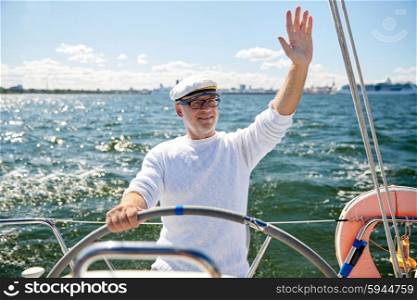 sailing, age, tourism, travel and people concept - happy senior man in captain hat on steering wheel and waving hand sail boat or yacht floating in sea