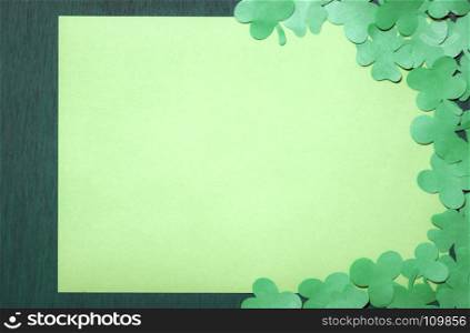 Saint Patrick greeting card design with lots of paper clovers surrounding an empty yellow paper sheet, displayed on a dark green wooden background.