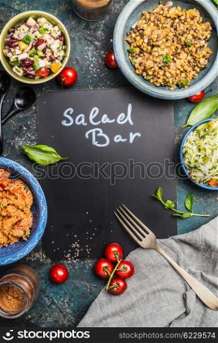 Salad bar with various healthy vegetarian salads dish, forg and black chalkboard, top view, frame. Inscription salad bar on blank chalkboard. Healthy food and vegetarian Eating concept