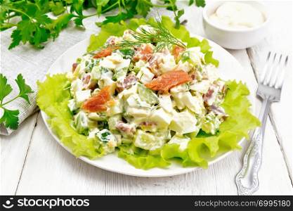 Salad from salmon, cucumber, eggs and avocado, dressed with mayonnaise on lettuce leaves in a plate, towel, dill, parsley and fork on a white wooden board background