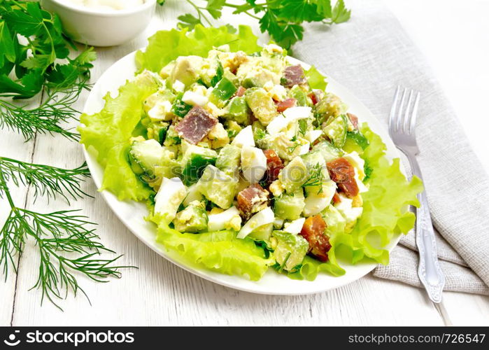 Salad from salmon, cucumber, eggs and avocado with mayonnaise on lettuce leaves in a plate, kitchen towel, dill, parsley and fork on a light wooden board background