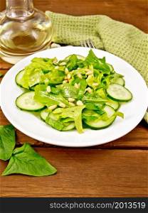 Salad from spinach, fresh cucumbers, rukkola salad, cedar nuts and spring onions, seasoned with vegetable oil on a plate, napkin and fork on a background of brown wooden board
