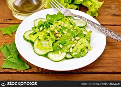 Salad from spinach, fresh cucumbers, rukkola salad, cedar nuts and spring onions, seasoned with vegetable oil and a fork on a plate on the background of a wooden board