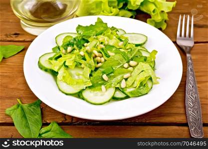 Salad from spinach, fresh cucumbers, rukkola salad, cedar nuts and spring onions, seasoned with vegetable oil on a plate, fork on a wooden plank background