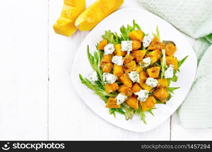 Salad of baked pumpkin, arugula with balls of salt cheese, seasoned with honey, grainy mustard, garlic and vegetable oil in a plate, napkin and fork on wooden board background from above