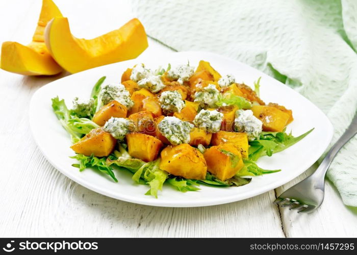 Salad of baked pumpkin, arugula with balls of salt cheese, seasoned with honey, mustard, garlic and vegetable oil in a plate, napkin and fork on light wooden board background