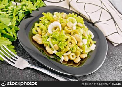 Salad of beans, olives, stalk celery, onion and lettuce, dressed with vegetable oil in a plate, towel and fork on wooden board background