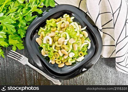 Salad of beans, olives, stalk celery, onion and lettuce, dressed with vegetable oil in a plate, napkin and fork on wooden board background from above