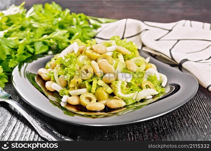Salad of beans, olives, stalk celery, onion and lettuce, dressed with vegetable oil in a plate, napkin and fork on dark wooden board background