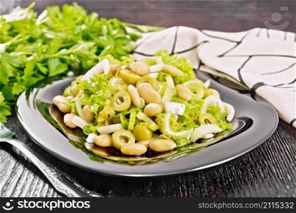 Salad of beans, olives, stalk celery, onion and lettuce, dressed with vegetable oil in a plate, napkin and fork on dark wooden board background