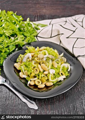 Salad of beans, olives, stalk celery, onion and lettuce, dressed with vegetable oil in a plate, napkin and fork on wooden board background